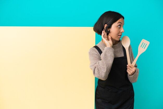 Jonge gemengd ras kok vrouw met een groot bord geïsoleerd op blauwe achtergrond luisteren naar iets door hand op het oor te leggen