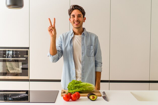 Jonge gemengd ras man bereidt een salade voor de lunch vrolijk en zorgeloos met een vredessymbool met vingers.