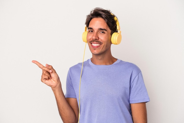 Jonge gemengd ras man luisteren naar muziek geïsoleerd op blauwe achtergrond glimlachend en opzij wijzend, iets tonen op lege ruimte.