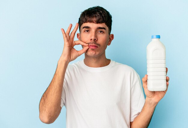 Jonge gemengd ras man met een fles melk geïsoleerd op blauwe achtergrond met vingers op lippen houden een geheim.