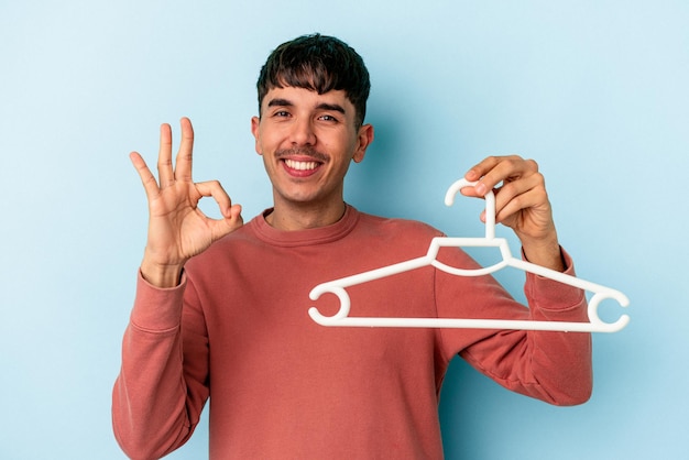 Jonge gemengd ras man met een hanger geïsoleerd op blauwe achtergrond vrolijk en zelfverzekerd weergegeven: ok gebaar.