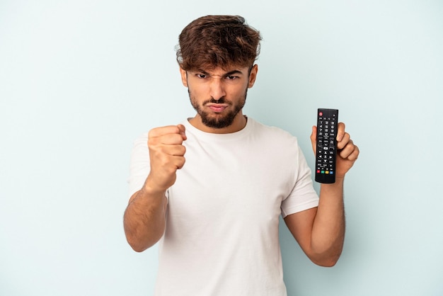 Jonge gemengd ras man met een tv-controller geïsoleerd op blauwe achtergrond met vuist naar camera, agressieve gezichtsuitdrukking.