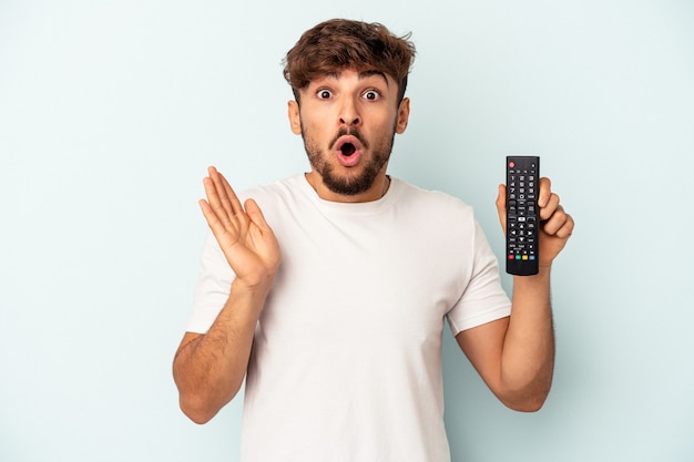 Jonge gemengd ras man met een tv-controller geïsoleerd op blauwe achtergrond verrast en geschokt.