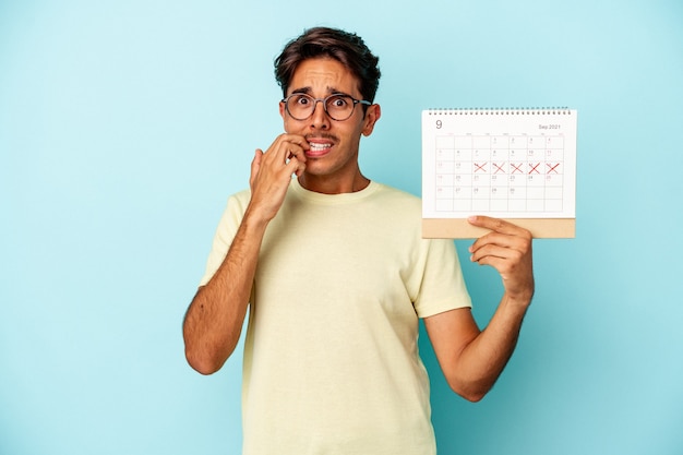 Jonge gemengd ras man met kalender geïsoleerd op blauwe achtergrond vingernagels bijten, nerveus en erg angstig.