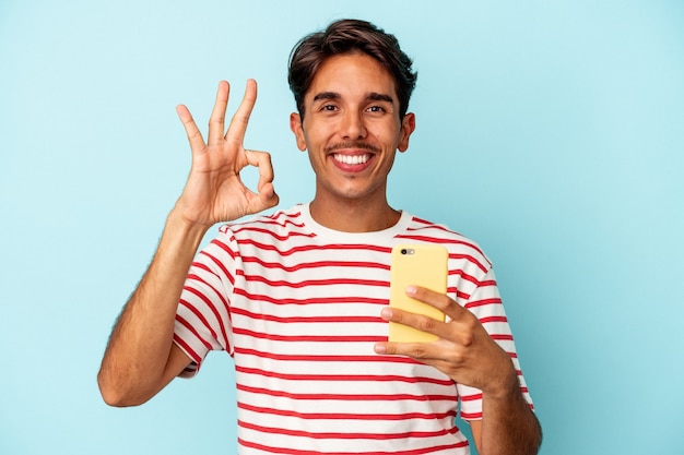 Jonge gemengd ras man met mobiele telefoon geïsoleerd op blauwe achtergrond vrolijk en zelfverzekerd weergegeven: ok gebaar.