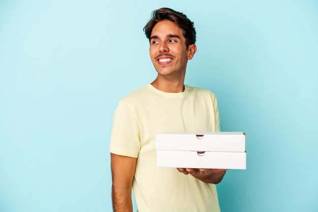 Jonge gemengd ras man met pizza's geïsoleerd op blauwe achtergrond kijkt opzij glimlachend, vrolijk en aangenaam.