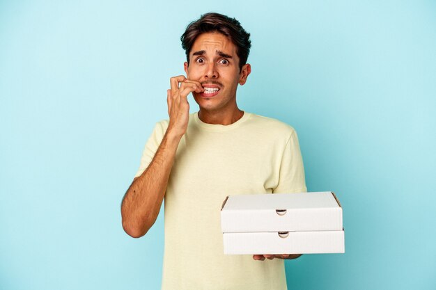 Jonge gemengd ras man met pizza's geïsoleerd op blauwe achtergrond vingernagels bijten, nerveus en erg angstig.