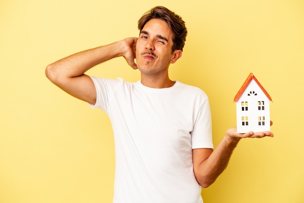 Jonge gemengd ras man met speelgoed huis geïsoleerd op gele achtergrond achterhoofd aan te raken, denken en een keuze maken.