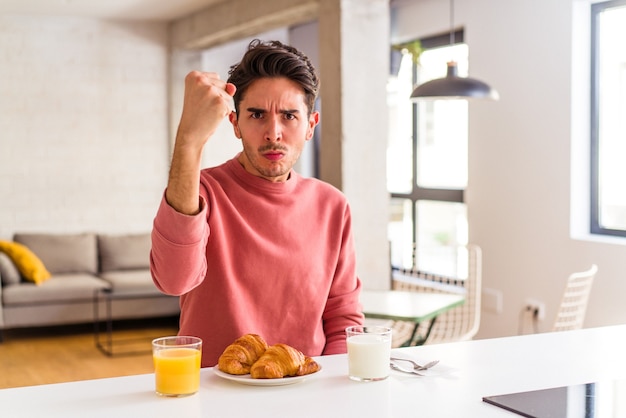 Jonge gemengd ras man ontbijten in een keuken op de ochtend met vuist naar camera, agressieve gezichtsuitdrukking.