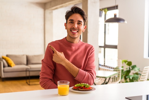 Jonge gemengd ras man ontbijten in zijn keuken glimlachend en opzij wijzend, iets laten zien op lege ruimte.