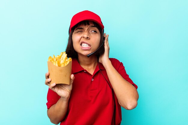 Jonge gemengd ras vrouw fastfood restaurant werknemer met frietjes geïsoleerd op blauwe achtergrond die oren met handen bedekt.