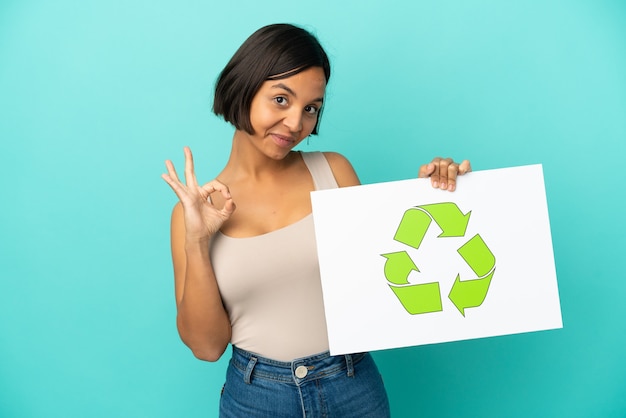 Jonge gemengd ras vrouw geïsoleerd op blauwe achtergrond met een bordje met recycle pictogram en een overwinning vieren