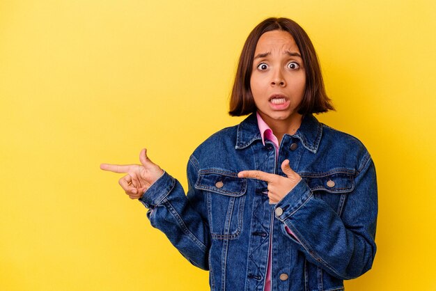 Jonge gemengd ras vrouw geïsoleerd op gele achtergrond geschokt wijzend met wijsvingers naar een kopieerruimte.