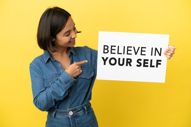 Jonge gemengd ras vrouw geïsoleerd op gele achtergrond met een bordje met tekst Believe In Your Self en wijst erop