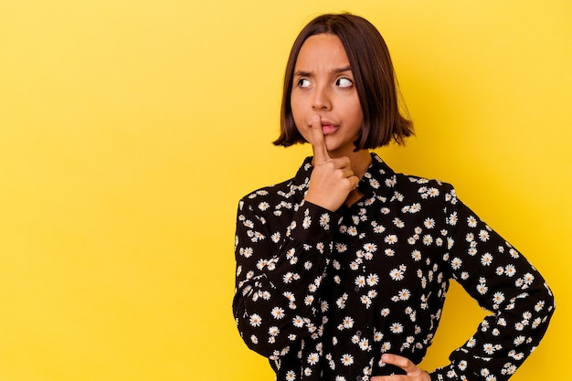 Jonge gemengd ras vrouw geïsoleerd op gele achtergrond zijwaarts kijkend met twijfelachtige en sceptische uitdrukking.