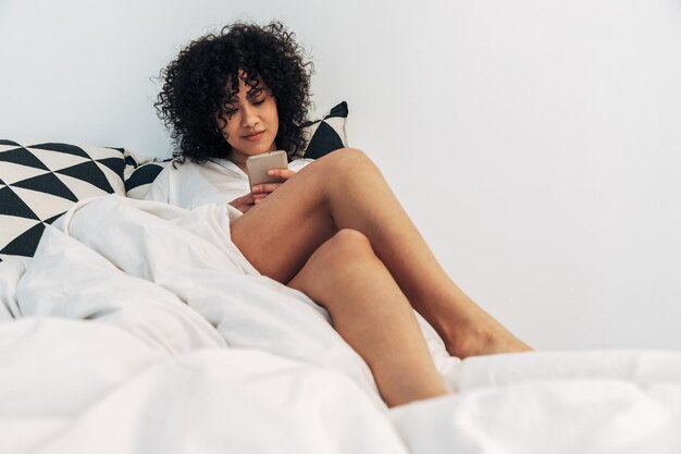 Jonge gemengd ras vrouw met behulp van mobiele telefoon in bed. Ruimte kopiëren. Lifestyle en sociale media concept.