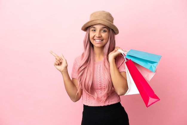 Jonge gemengd ras vrouw met boodschappentas geïsoleerd op roze achtergrond wijzende vinger naar de zijkant