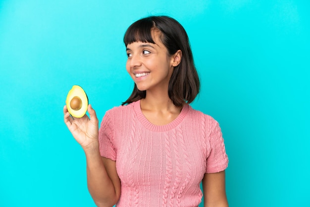 Jonge gemengd ras vrouw met een avocado geïsoleerd op een blauwe achtergrond op zoek naar kant