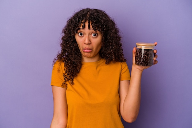Jonge gemengd ras vrouw met een fles koffiebonen geïsoleerd op paarse achtergrond haalt schouders op en open ogen verward.