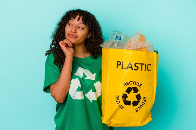 Jonge gemengd ras vrouw met een gerecyclede plastic zak geïsoleerd op een blauwe achtergrond zijwaarts kijkend met twijfelachtige en sceptische uitdrukking.