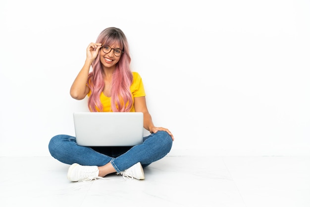 Jonge gemengd ras vrouw met een laptop met roze haren zittend op de vloer geïsoleerd op een witte achtergrond met een bril en happy