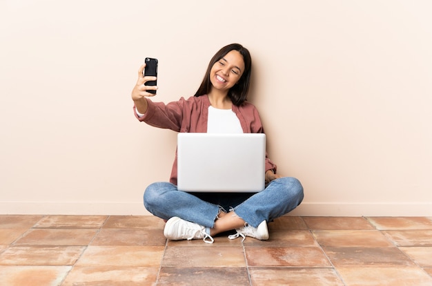 Jonge gemengd ras vrouw met een laptop zittend op de vloer een selfie maken