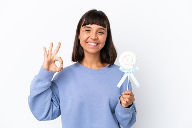 Jonge gemengd ras vrouw met een lolly geïsoleerd op een witte achtergrond met ok teken met vingers