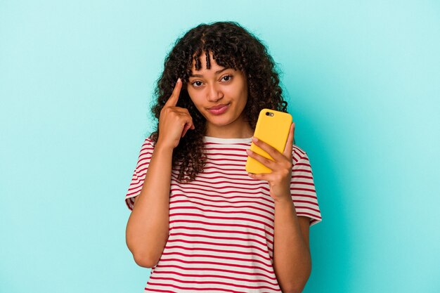 Jonge gemengd ras vrouw met een mobiele telefoon geïsoleerd op blauwe achtergrond wijzende tempel met vinger, denken, gericht op een taak.