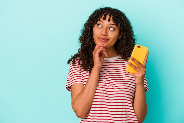 Jonge gemengd ras vrouw met een mobiele telefoon geïsoleerd op blauwe achtergrond zijwaarts kijkend met twijfelachtige en sceptische uitdrukking.