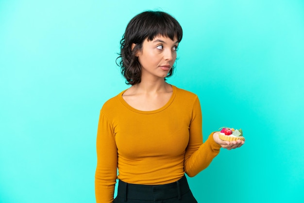 Jonge gemengd ras vrouw met een taartje geïsoleerd op een blauwe achtergrond op zoek naar de kant