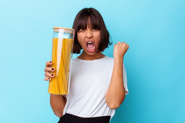 Foto jonge gemengd ras vrouw met pasta pot geïsoleerd op blauwe achtergrond verhogen vuist na een overwinning, winnaar concept.