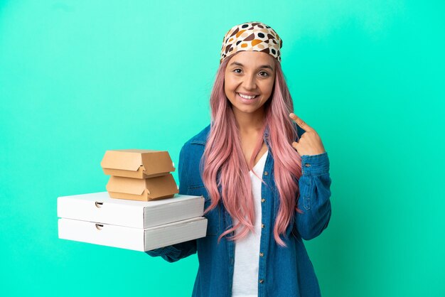 Jonge gemengd ras vrouw met pizza's en hamburgers geïsoleerd op een groene achtergrond met een duim omhoog gebaar