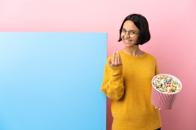 Jonge gemengd ras vrouw met popcorns met een grote banner over geïsoleerde achtergrond uitnodigend om met de hand te komen. Blij dat je gekomen bent