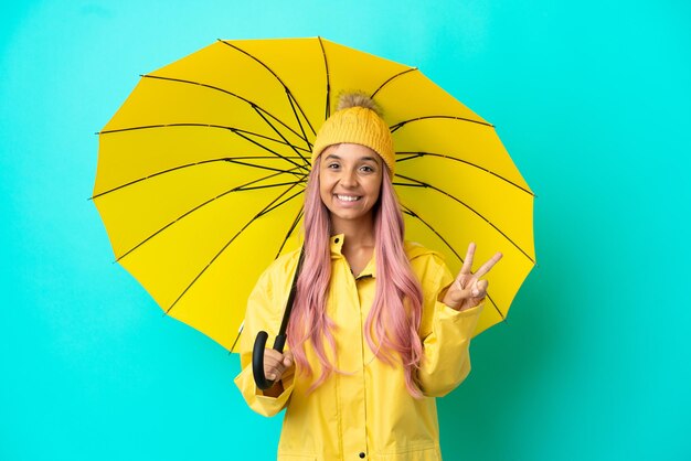 Jonge gemengd ras vrouw met regendichte jas en paraplu glimlachend en overwinningsteken tonen