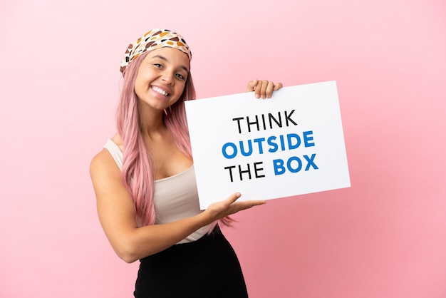 Jonge gemengd ras vrouw met roze haar geïsoleerd op roze achtergrond met een bordje met tekst Think Outside The Box met gelukkige uitdrukking