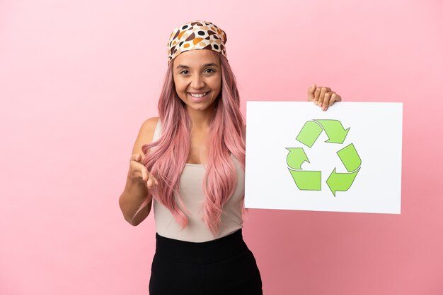 Jonge gemengd ras vrouw met roze haar geïsoleerd op roze achtergrond met een plakkaat met recycle icoon maken van een deal