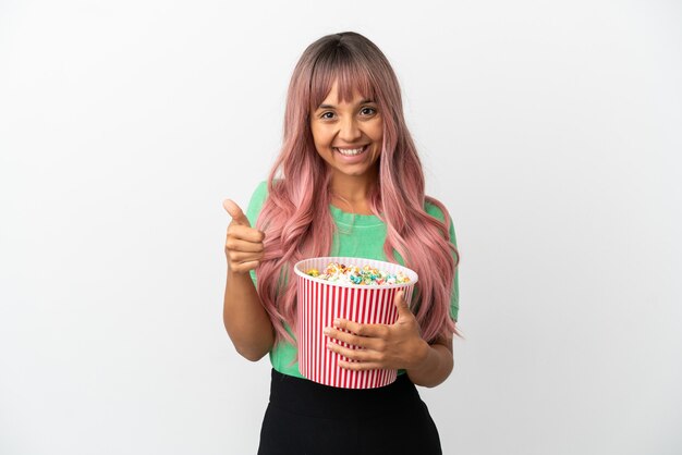 Jonge gemengd ras vrouw met roze haren eten popcorn geïsoleerd op een witte achtergrond met duimen omhoog omdat er iets goeds is gebeurd