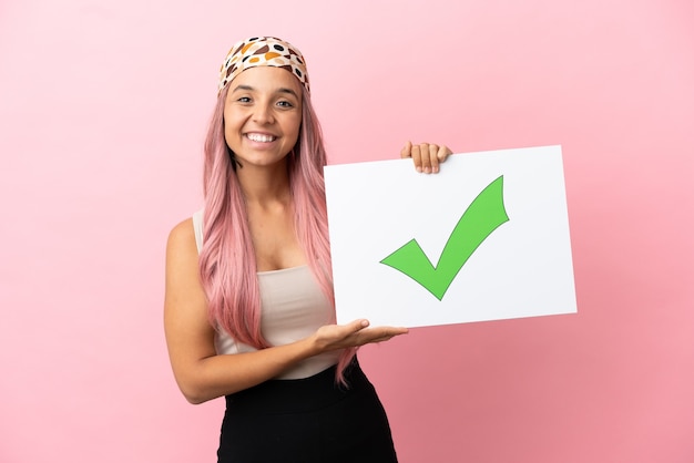 Jonge gemengd ras vrouw met roze haren geïsoleerd op roze achtergrond met een bordje met tekst Groen vinkje pictogram met gelukkige expressie