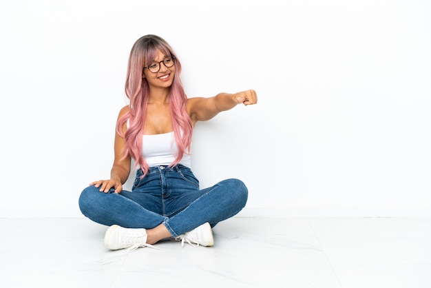 Jonge gemengd ras vrouw met roze haren zittend op de vloer geïsoleerd op een witte achtergrond met een duim omhoog gebaar