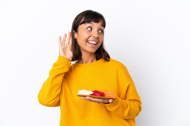 Jonge gemengd ras vrouw met sashimi geïsoleerd op een witte achtergrond luisteren naar iets door hand op het oor te leggen