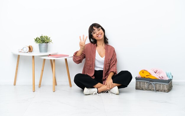 Jonge gemengd ras vrouw vouwen kleren zittend op de vloer geïsoleerd op een witte achtergrond gelukkig en tellen drie met vingers