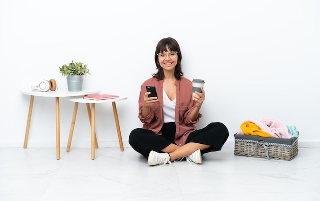 Jonge gemengd ras vrouw vouwen kleren zittend op de vloer geïsoleerd op een witte achtergrond met koffie om mee te nemen en een mobiel