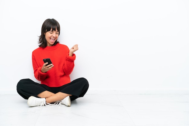 Jonge gemengd ras vrouw zittend op de vloer geïsoleerd op een witte achtergrond met telefoon in overwinningspositie