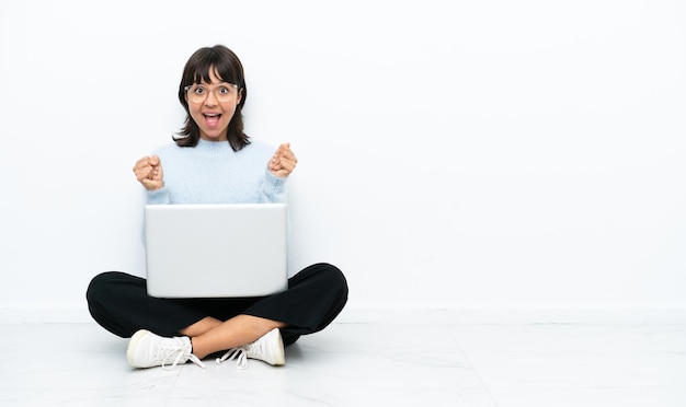 Jonge gemengd ras vrouw zittend op de vloer met laptop geïsoleerd op een witte achtergrond vieren een overwinning in winnaar positie