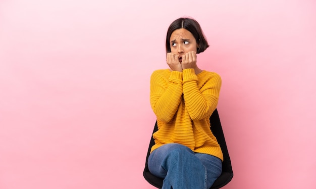 Jonge gemengd ras vrouw zittend op een stoel geïsoleerd op roze achtergrond nerveus en bang handen op de mond