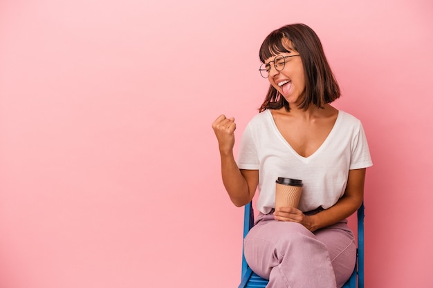 Jonge gemengd ras vrouw zittend op een stoel met een kopje koffie geïsoleerd op roze achtergrond verhogen vuist na een overwinning, winnaar concept.