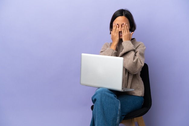 Jonge gemengd ras vrouw zittend op een stoel met laptop geïsoleerd op een paarse achtergrond die ogen bedekt door handen