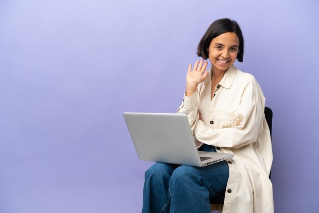 Jonge gemengd ras vrouw zittend op een stoel met laptop geïsoleerd op paarse achtergrond groeten met hand met gelukkige uitdrukking