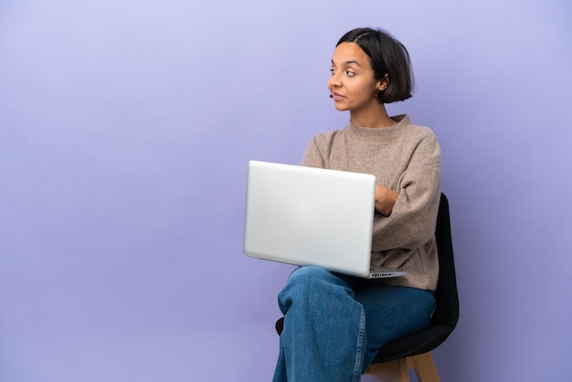 Jonge gemengd ras vrouw zittend op een stoel met laptop geïsoleerd op paarse achtergrond in laterale positie