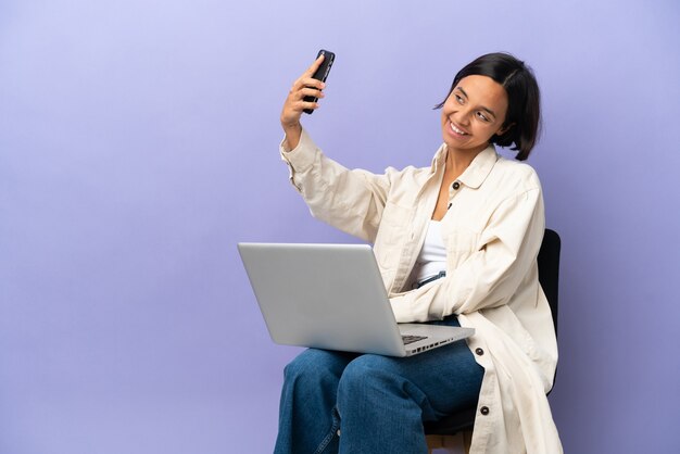 Jonge gemengd ras vrouw zittend op een stoel met laptop geïsoleerd op paarse achtergrond maken een selfie